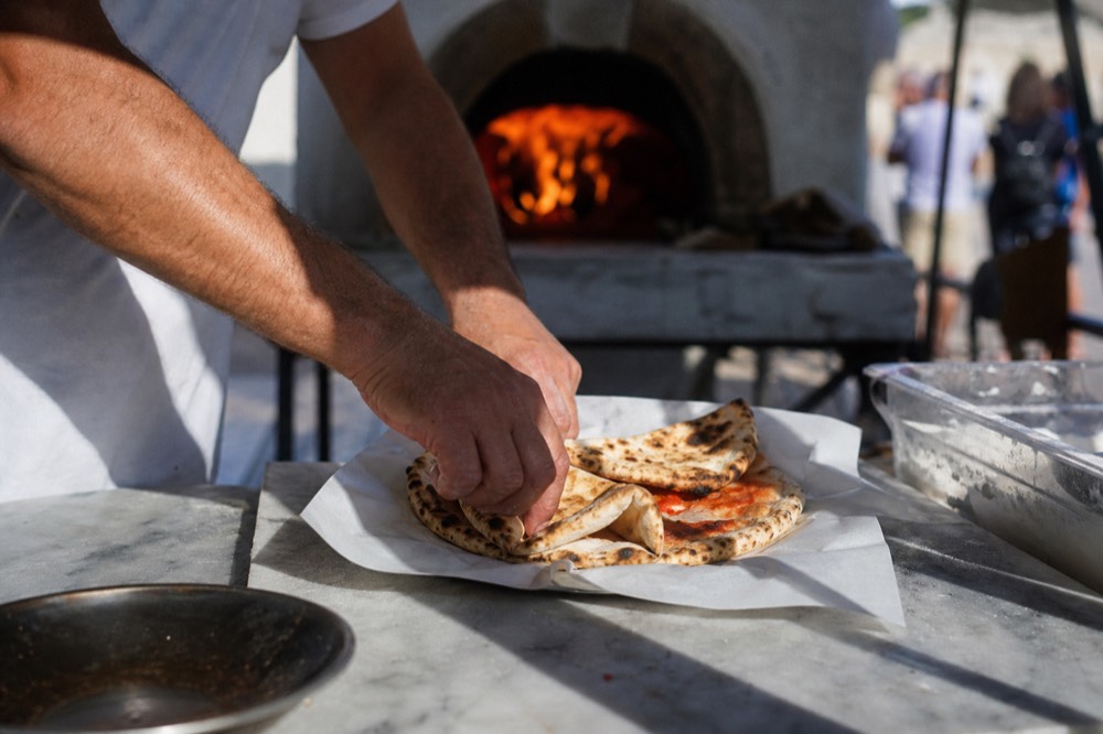 Baker's hands folding a hot pizza into quarters over greaseproof paper at a street counter, wood oven glowing orange in the background