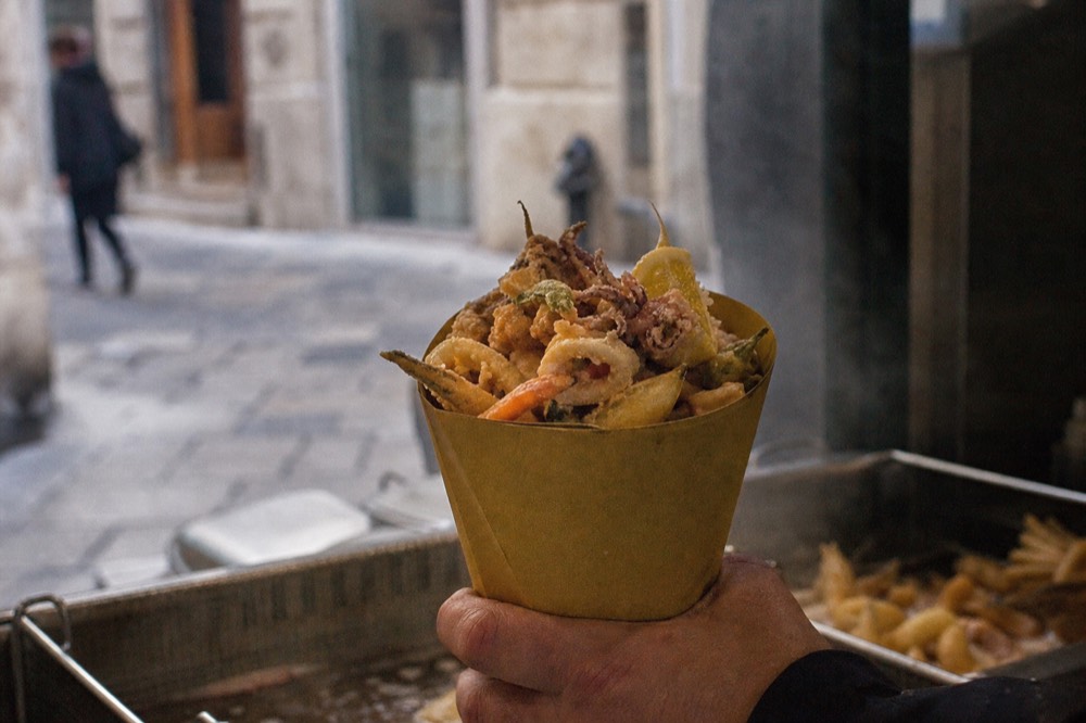 Paper cone of golden fried seafood and vegetables held over a fryer basket, oil steam visible, tiled shopfront blurred behind