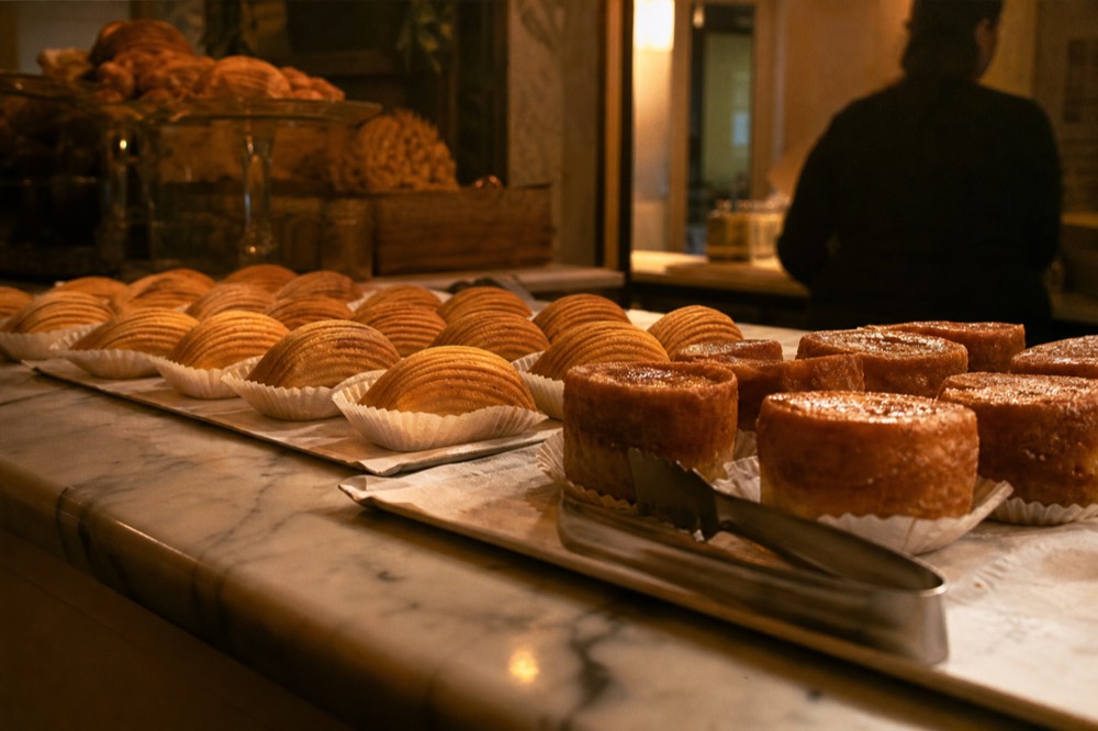 Marble pastry counter lined with ribbed shell pastries and rum-soaked sponge cakes under warm light, tongs resting on a paper-lined tray