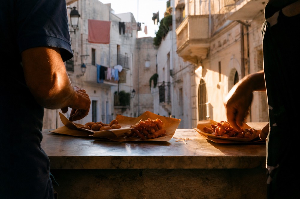 Small group standing around a marble street counter tasting fried snacks from paper, narrow sunlit alley with laundry lines overhead